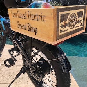 May include: An electric bicycle with a wooden cargo box attached to the rear. The box is branded with "East Coast Electric Speed Shop" in a vintage style. The bike is black with blue accents and has a helmet inside the box.