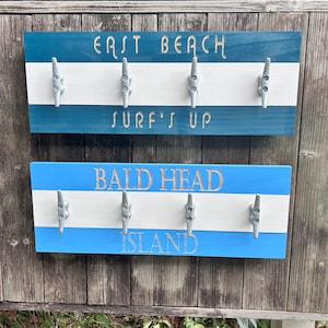 May include: Two wooden signs with nautical rope hooks. The top sign is blue and white with the text "East Beach" and "Surf's Up". The bottom sign is blue and white with the text "Bald Head Island".