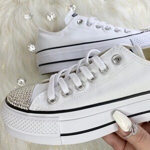 May include: White platform Converse sneakers with a black trim and a row of rhinestones on the toe.