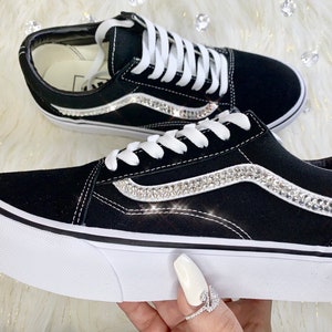 K&ouml;nnte beinhalten: Schwarze Canvas-Sneaker mit wei&szlig;en Schn&uuml;rsenkeln und wei&szlig;er Gummisohle. Die Seite des Schuhs hat einen wei&szlig;en Streifen mit einer Reihe von klaren Strasssteinen.