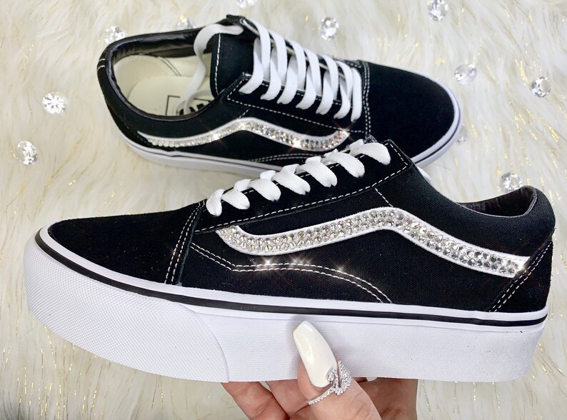 K&ouml;nnte beinhalten: Schwarze Canvas-Sneaker mit wei&szlig;en Schn&uuml;rsenkeln und wei&szlig;er Gummisohle. Die Seite des Schuhs hat einen wei&szlig;en Streifen mit einer Reihe von klaren Strasssteinen.