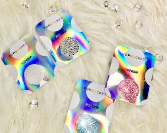 May include: Four PopSocket PopGrip phone grips with different designs. The grips are on a white fuzzy surface. The designs are clear with white, silver, pink, and blue glitter.