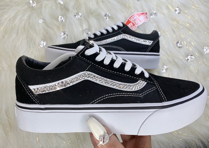 K&ouml;nnte beinhalten: Schwarze Vans-Sneaker aus Canvas mit einer wei&szlig;en Plateau-Sohle und einer Reihe von Strasssteinen auf dem seitlichen Streifen. Das Vans-Logo ist an der Seite des Schuhs sichtbar.