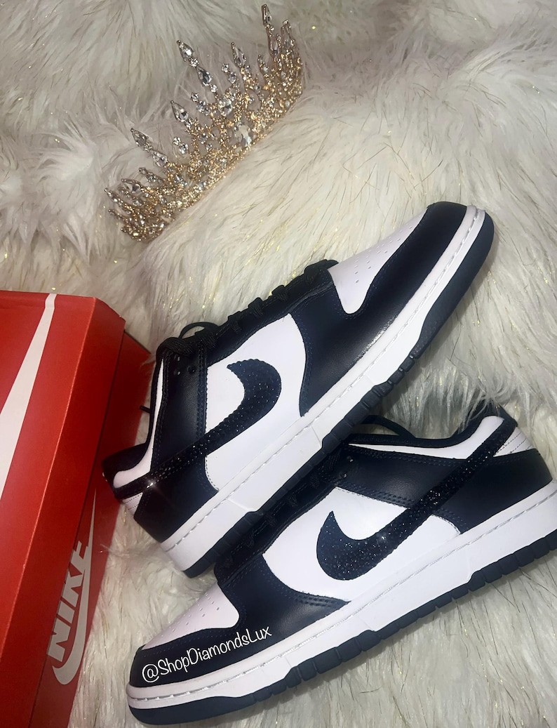 Peut inclure: Des baskets basses Nike noires et blanches avec un swoosh noir scintillant. Les baskets sont sur une surface moelleuse blanche avec une couronne dor&eacute;e.