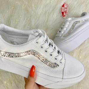 May include: White sneakers with a platform sole, embellished with sparkling rhinestones along the side stripe. The shoes have white laces and a classic design, perfect for casual wear. A Vans logo sticker is visible.