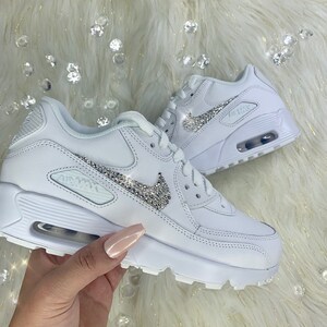 May include: White leather sneakers with a white swoosh logo adorned with silver rhinestones. The sneakers have a white rubber sole and a white air max logo on the side.