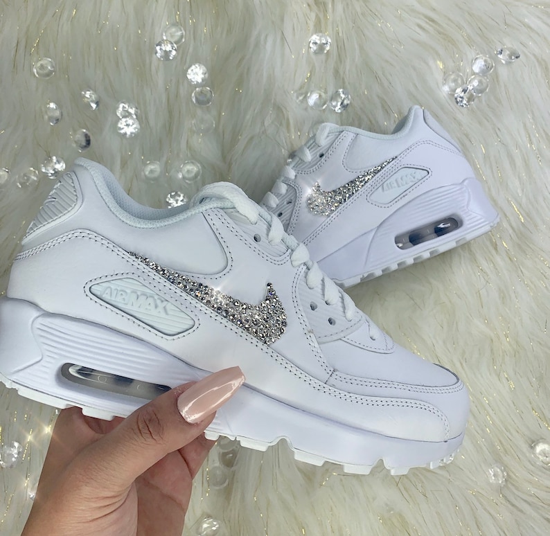 May include: White leather sneakers with a white swoosh logo adorned with silver rhinestones. The sneakers have a white rubber sole and a white air max logo on the side.