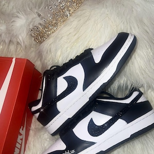 Peut inclure: Des baskets basses Nike noires et blanches avec un swoosh noir scintillant. Les baskets sont sur une surface moelleuse blanche avec une couronne dor&eacute;e.