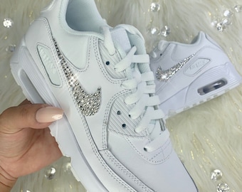May include: White leather sneakers with a white swoosh and a row of rhinestones on the side. The sneakers have a white sole and a white lace.