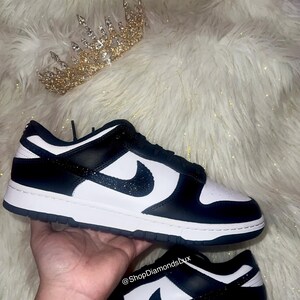 Peut inclure: Des baskets basses noires et blanches avec un swoosh noir et des accents de paillettes noires. Les baskets sont sur une surface en fourrure blanche. Le texte "@ShopDiamondsLux" est visible sur l'image.