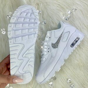 May include: White leather Nike Air Max 90 sneakers with a white swoosh decorated with rhinestones. The sneakers have a white rubber sole.