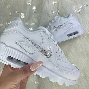 May include: White leather sneakers with a silver rhinestone Nike swoosh logo. The sneakers have a white rubber sole and the text "AIR MAX" is printed on the side.