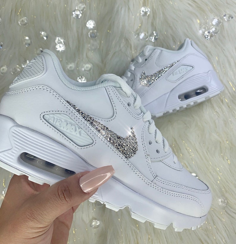 May include: White leather sneakers with a silver rhinestone Nike swoosh logo. The sneakers have a white rubber sole and the text "AIR MAX" is printed on the side.