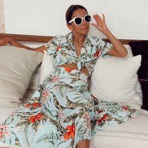 May include: A woman wearing a light blue tropical print two-piece outfit with a white button-down shirt and a matching skirt. She is wearing white round sunglasses and sitting on a white sofa with cushions.