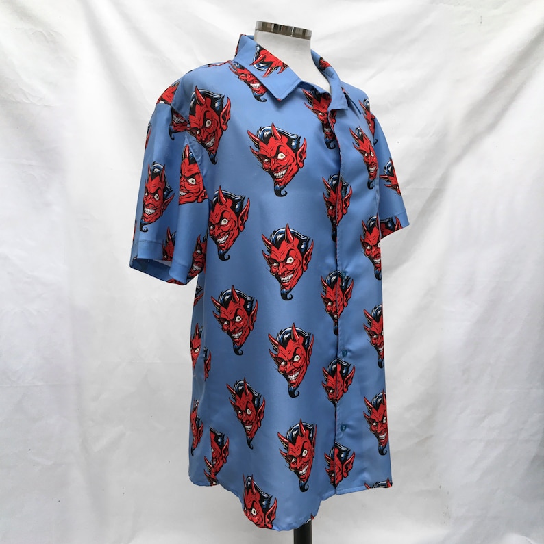 Pode incluir: Camisa azul clara com bot&otilde;es com um padr&atilde;o repetido de rostos de dem&ocirc;nios vermelhos. A camisa de manga curta &eacute; exibida em um manequim em um fundo branco. Os rostos dos dem&ocirc;nios t&ecirc;m chifres e um sorriso sinistro.