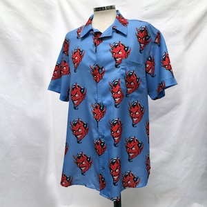 Pode incluir: Camisa azul clara de manga curta com um padr&atilde;o repetido de rostos de dem&ocirc;nios vermelhos com chifres e barbas pretas. A camisa tem gola e um bolso no peito. A camisa &eacute; exibida em um manequim.