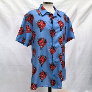 Pode incluir: Camisa azul clara de manga curta com um padr&atilde;o repetido de rostos de dem&oacute;nios vermelhos. A camisa tem uma gola cl&aacute;ssica e um bolso no peito. Os rostos dos dem&oacute;nios t&ecirc;m chifres vermelhos e um sorriso malicioso.