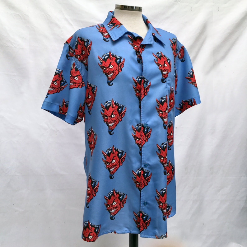 Pode incluir: Camisa azul clara de manga curta com um padr&atilde;o repetido de rostos de dem&oacute;nios vermelhos. A camisa tem uma gola cl&aacute;ssica e um bolso no peito. Os rostos dos dem&oacute;nios t&ecirc;m chifres vermelhos e um sorriso malicioso.