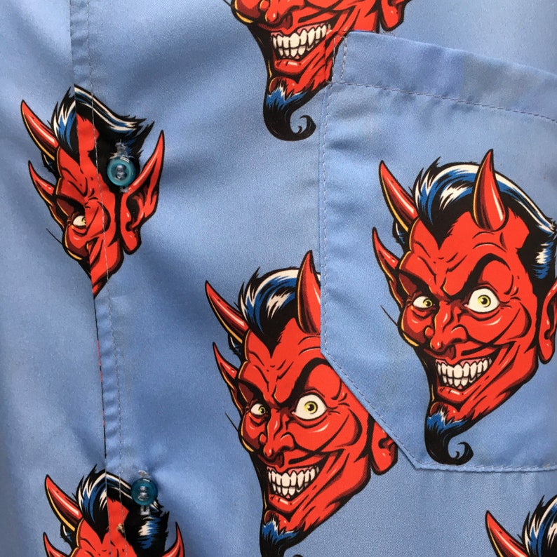 Pode incluir: Camisa azul clara com bot&otilde;es com um padr&atilde;o repetido de rostos de dem&ocirc;nios vermelhos com chifres, cabelo azul e dentes brancos. A camisa tem um bolso no lado esquerdo e bot&otilde;es azuis.