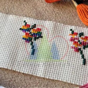 Op de afbeelding: Een kruissteek bloemenontwerp op witte stof, met drie kleurrijke bloemboeketten met groene stelen. Borduurgaren in verschillende kleuren, waaronder oranje, roze en wit, omringt de stof.