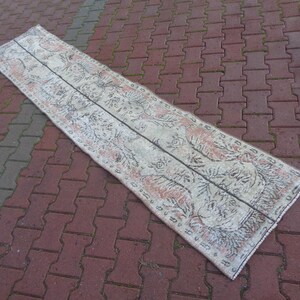 May include: A white and cream runner rug with a floral pattern in black and pink. The rug has a distressed look and is made of a soft, woven material.