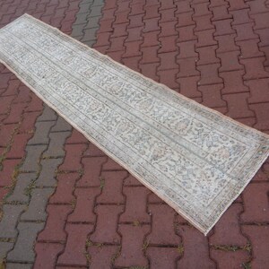 May include: A long, rectangular runner rug with a white background and a delicate floral pattern in shades of gray and pink. The rug has a fringed edge.