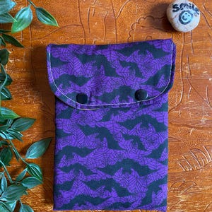 May include: A purple and black fabric pouch with a flap closure, featuring a bat and spiderweb design. The pouch is fastened with two black buttons. The background is a wooden surface with green leaves.