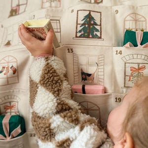 May include: Advent calendar with numbered window pockets, each featuring a festive illustration. A child's arm reaches for a small box. The calendar is made of beige fabric with brown and white accents. The box has the word "MOUSE" on it.