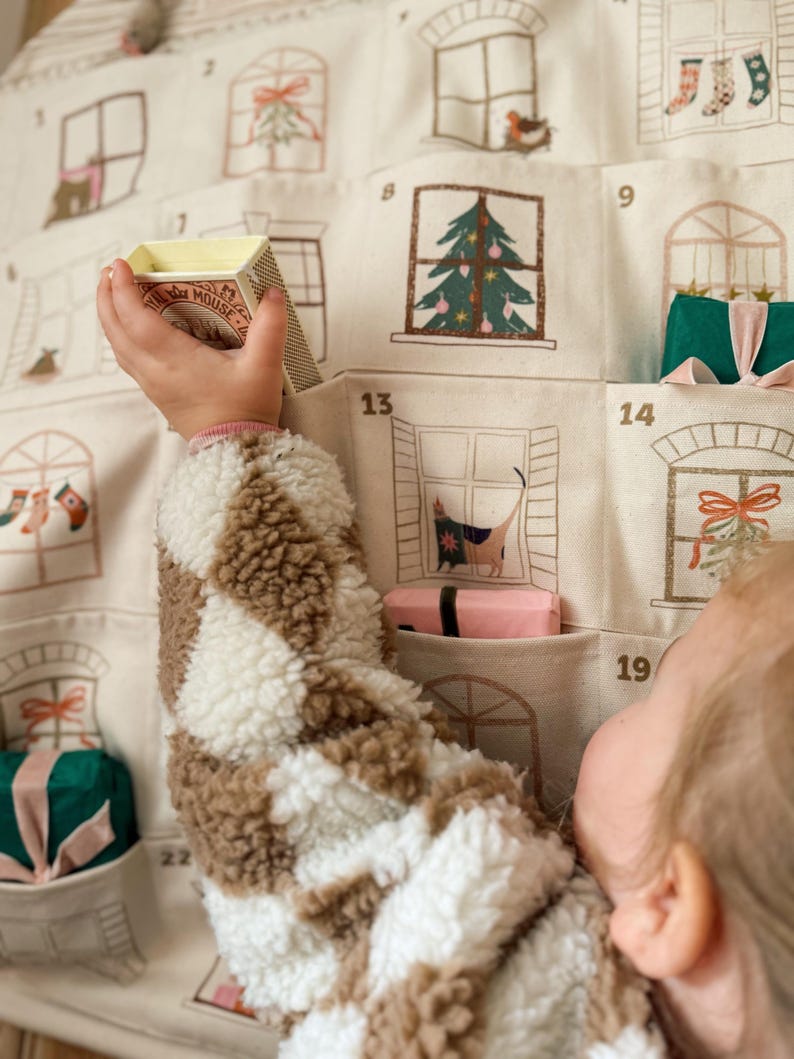 May include: Advent calendar with numbered window pockets, each featuring a festive illustration. A child's arm reaches for a small box. The calendar is made of beige fabric with brown and white accents. The box has the word "MOUSE" on it.
