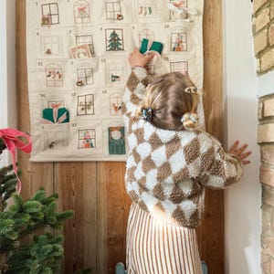 May include: A beige fabric advent calendar shaped like a house, with embroidered windows and pockets. A child in a brown and white checkered jacket and striped pants reaches for a gift. A Christmas tree is in the foreground.