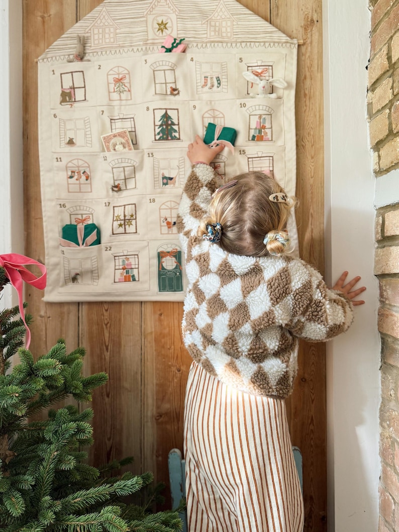 May include: A beige fabric advent calendar shaped like a house, with embroidered windows and pockets. A child in a brown and white checkered jacket and striped pants reaches for a gift. A Christmas tree is in the foreground.