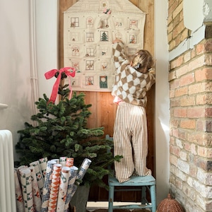 May include: A Christmas advent calendar shaped like a house, with small windows, hangs on a wooden door. A small child in a checkered sweater and striped pants reaches for the calendar. A small Christmas tree and wrapped gifts are in the foreground.