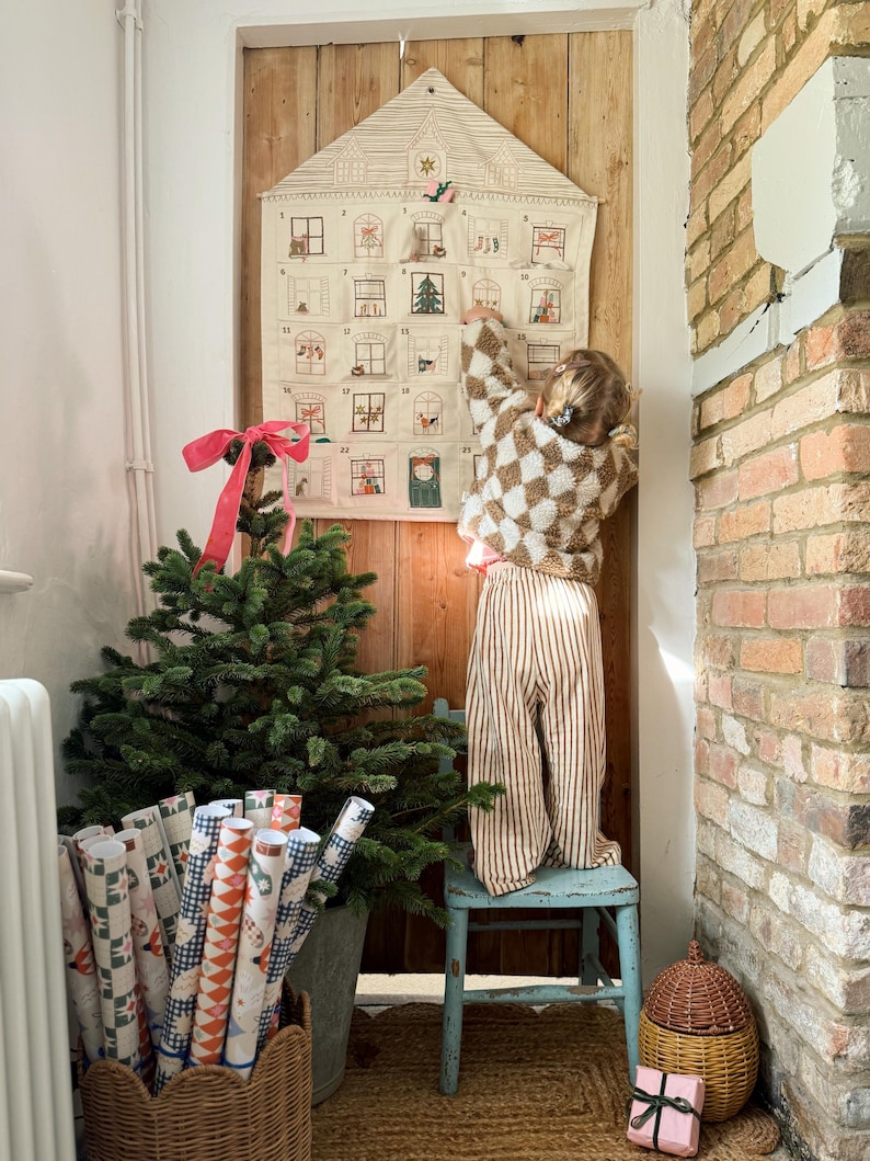 May include: A Christmas advent calendar shaped like a house, with small windows, hangs on a wooden door. A small child in a checkered sweater and striped pants reaches for the calendar. A small Christmas tree and wrapped gifts are in the foreground.