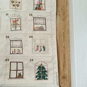 May include: A cream-colored advent calendar with embroidered window designs. Each window depicts a festive scene, including gifts, stars, and a Christmas tree. The calendar is mounted on a wooden dowel and features the text "KNOTTY KNOTTY".
