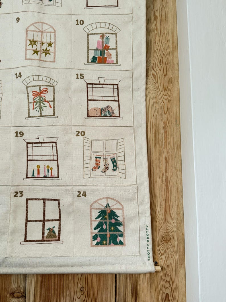 May include: A cream-colored advent calendar with embroidered window designs. Each window depicts a festive scene, including gifts, stars, and a Christmas tree. The calendar is mounted on a wooden dowel and features the text "KNOTTY KNOTTY".