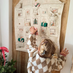 May include: A beige fabric advent calendar shaped like a house, with embroidered windows and pockets holding small gifts. A child in a brown and white checkered jacket reaches for a gift. The calendar hangs on a wooden wall.