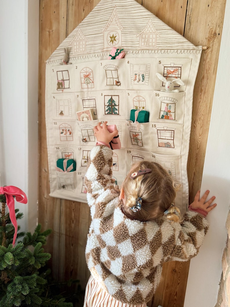 May include: A beige fabric advent calendar shaped like a house, with embroidered windows and pockets holding small gifts. A child in a brown and white checkered jacket reaches for a gift. The calendar hangs on a wooden wall.