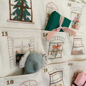 May include: A cream-colored fabric advent calendar with pockets, each numbered and decorated with window illustrations. A green gift box with a pink ribbon sits in one pocket, and a blue elephant plush toy peeks out of another. The calendar features Christmas-themed drawings.