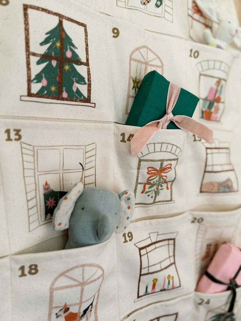 May include: A cream-colored fabric advent calendar with pockets, each numbered and decorated with window illustrations. A green gift box with a pink ribbon sits in one pocket, and a blue elephant plush toy peeks out of another. The calendar features Christmas-themed drawings.