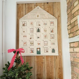May include: A fabric advent calendar shaped like a house, with 24 numbered windows, each depicting a festive scene. The calendar is cream-colored and hangs on a wooden wall. A small Christmas tree with a pink bow sits in front of it.