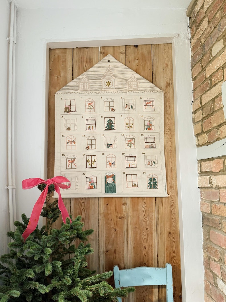 May include: A fabric advent calendar shaped like a house, with 24 numbered windows, each depicting a festive scene. The calendar is cream-colored and hangs on a wooden wall. A small Christmas tree with a pink bow sits in front of it.