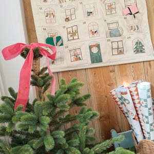 May include: A Christmas advent calendar with embroidered window designs, featuring small pockets for treats. A small Christmas tree with a pink ribbon is in the foreground. Rolls of patterned wrapping paper are in a wicker basket.