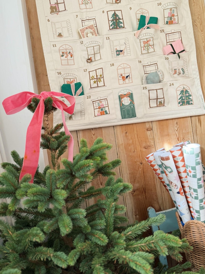 May include: A Christmas advent calendar with embroidered window designs, featuring small pockets for treats. A small Christmas tree with a pink ribbon is in the foreground. Rolls of patterned wrapping paper are in a wicker basket.