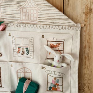 May include: A cream-colored fabric advent calendar with house and window illustrations. A white stuffed bunny is in one pocket. Other pockets contain a wrapped green gift and Christmas stockings.