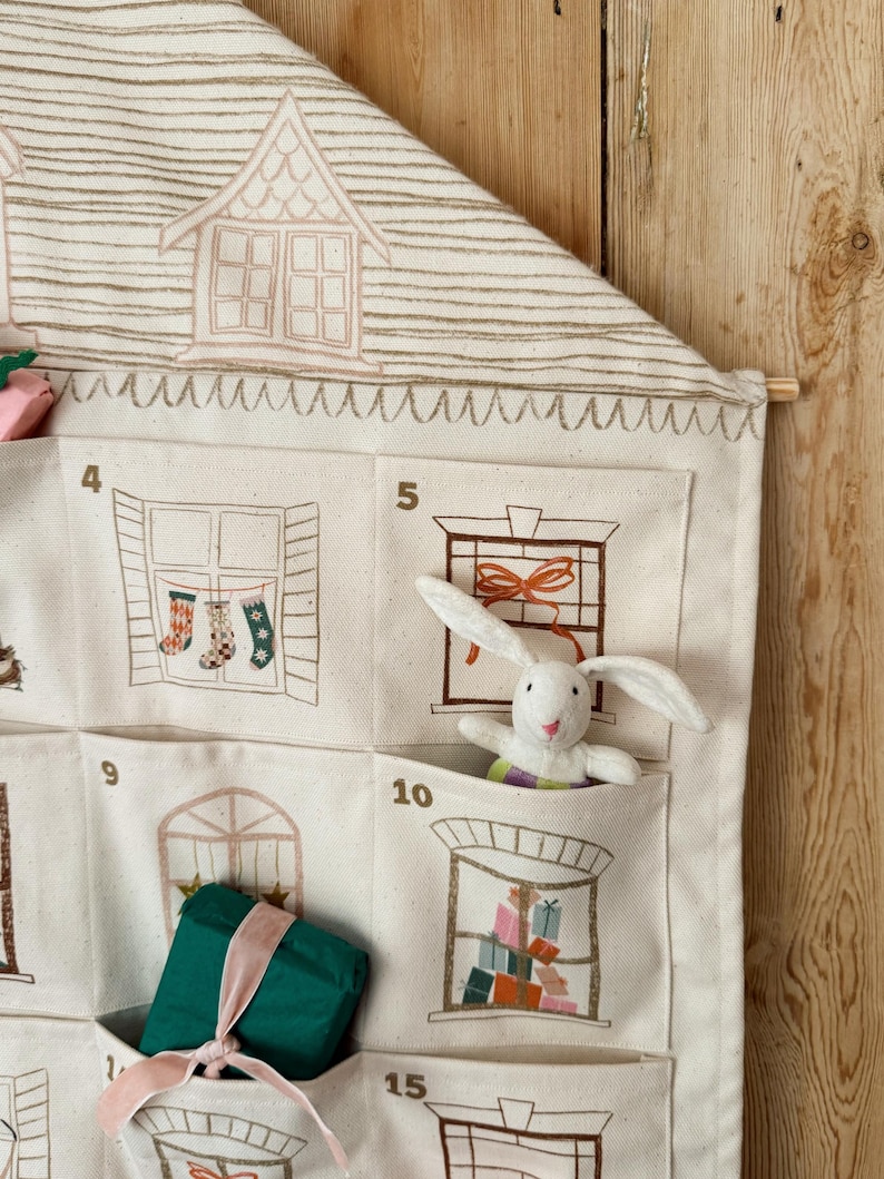 May include: A cream-colored fabric advent calendar with house and window illustrations. A white stuffed bunny is in one pocket. Other pockets contain a wrapped green gift and Christmas stockings.