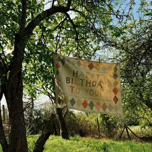 May include: A rectangular banner with the text "HAPPY BIRTHDAY TO YOU!" hangs from a clothesline in a sunny outdoor setting. The banner has a cream background with colorful diamond patterns and a decorative border.