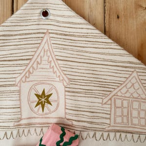 May include: Cream-colored fabric advent calendar with a house design in light brown. The calendar has pockets numbered 3 and 4, and a pink package with green trim is in one of the pockets. The top has a metal grommet for hanging.