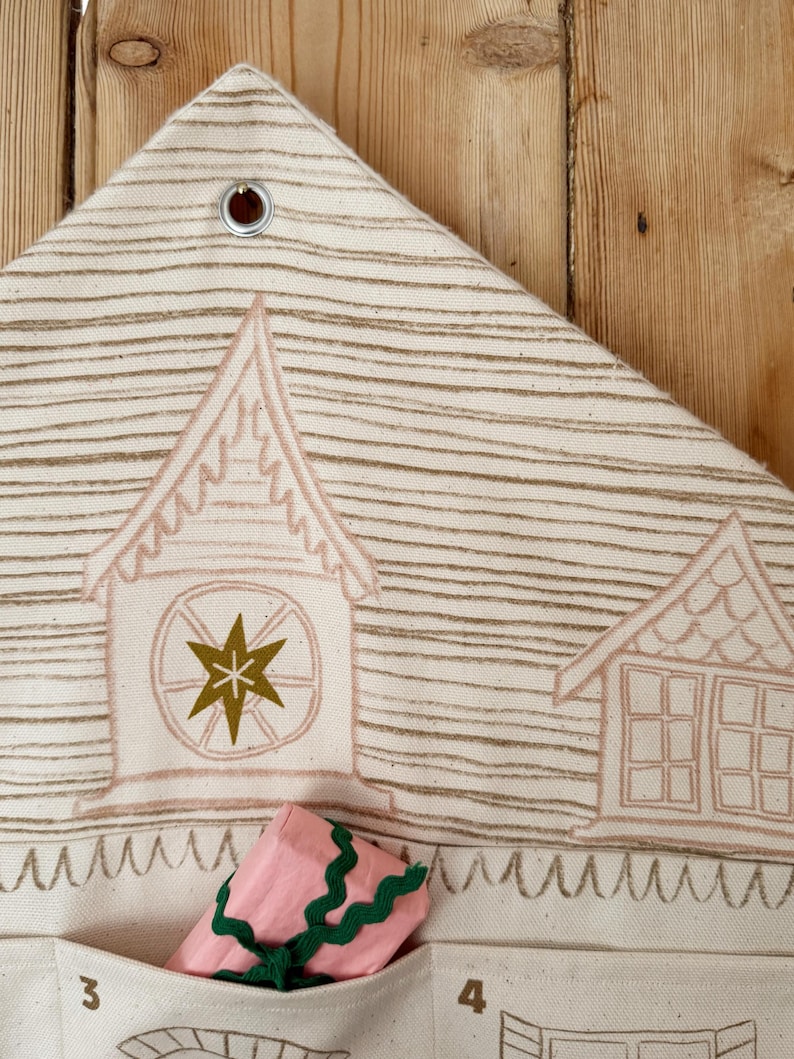 May include: Cream-colored fabric advent calendar with a house design in light brown. The calendar has pockets numbered 3 and 4, and a pink package with green trim is in one of the pockets. The top has a metal grommet for hanging.
