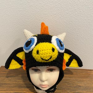 May include: A black crocheted hat featuring a yellow dragon face, large blue and white eyes, two white horns, an orange crest, and yellow and black wings on the sides. The hat has a chin strap and is displayed on a mannequin head.