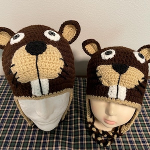 以下が含まれることがあります： 2 つの茶色のビーバーの形をしたかぎ針編みの帽子。帽子はベージュの顔、白い歯、黒い鼻、白い目です。帽子は白いマネキンの頭と大きな形の上にあります。帽子には耳あてが付いています。